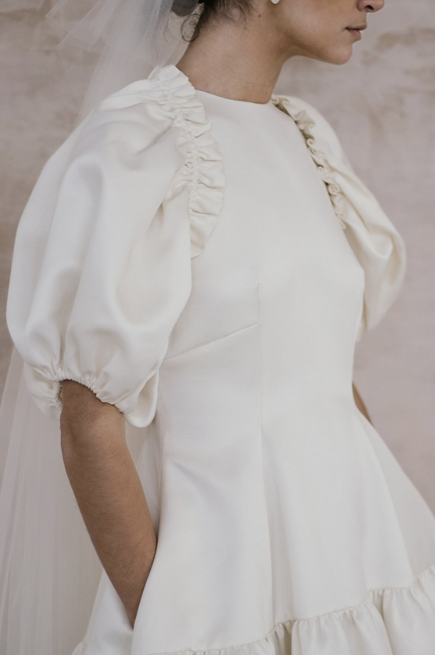 A close-up shot of a model wearing a dress with puffy sleeves, pleated at the shoulder and a simple white dress with a high neck under it